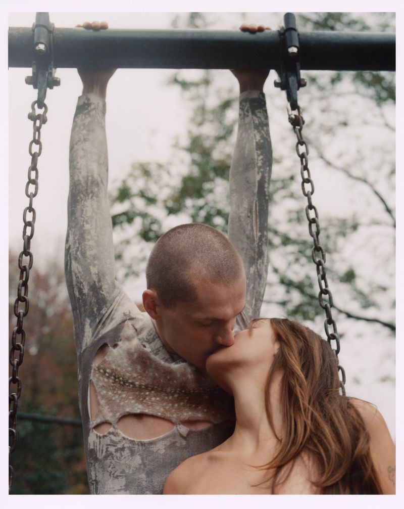 A couple embracing and kissing on a playground, the man hanging from a bar and the woman standing beneath him, photographed outdoors.