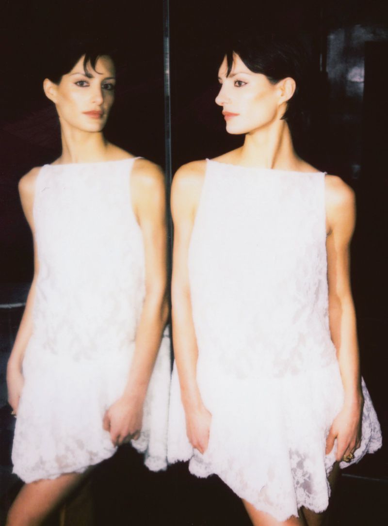 A woman with dark hair poses in a white dress, reflected in a mirror with a dark background in a fashion-style photograph.