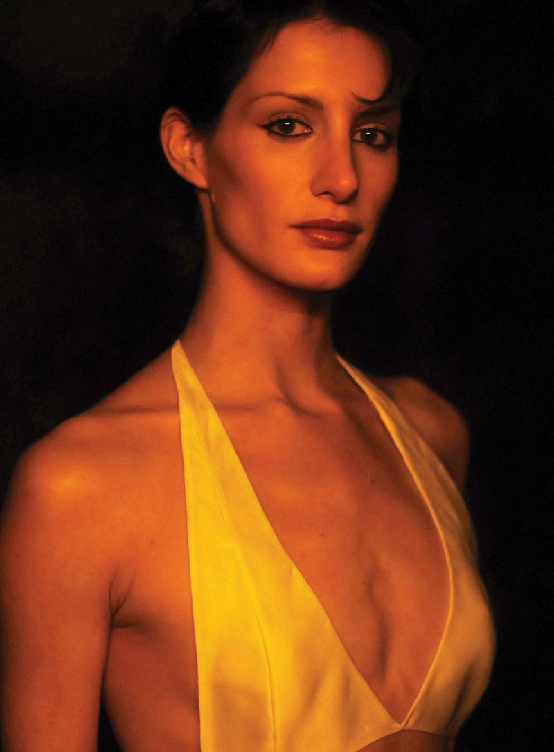 Portrait of a woman with dark hair and neutral expression wearing a yellow halter top against a dark background in a studio setting.