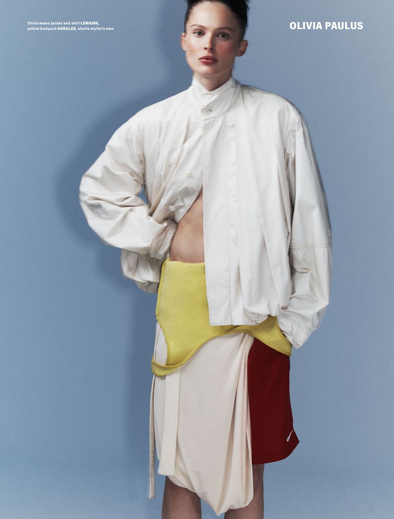 Fashion editorial image of a woman with short hair wearing a beige jacket, yellow top, and layered skirt on a light blue background.