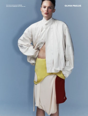 Fashion editorial image of a woman with short hair wearing a beige jacket, yellow top, and layered skirt on a light blue background.