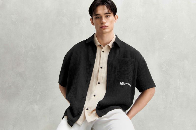A young adult male poses indoors wearing a black open-front shirt with logo over a tan button-down shirt and white pants.