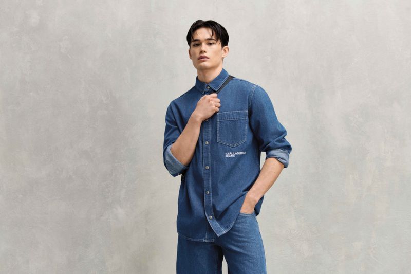 Asian male model poses in a blue denim shirt and jeans against a neutral background, showcasing a casual fashion style.