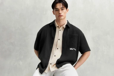A young male model wearing an unbuttoned black shirt over a light button-down shirt against a textured grey background.