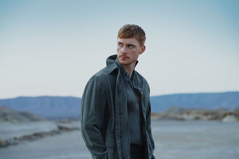 Adult man with red hair poses in a gray jacket and shirt, standing outdoors against a pale sky and distant mountains in a stylish portrait.
