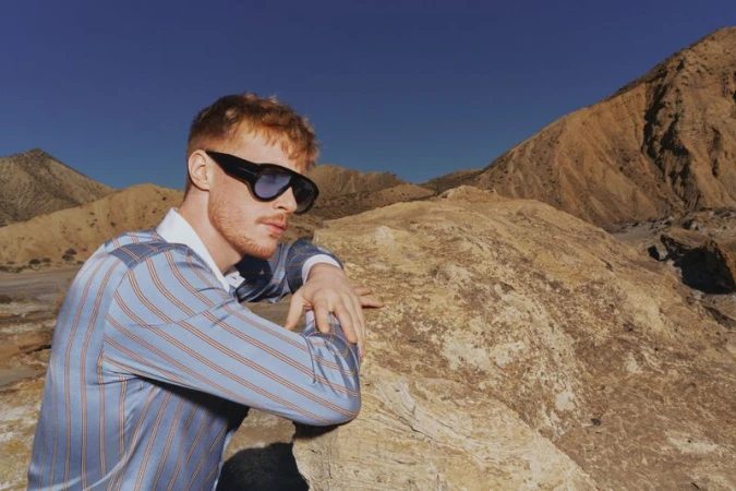 Adult male with red hair and sunglasses poses in a striped shirt, leaning on a rock formation with mountains and clear blue sky.