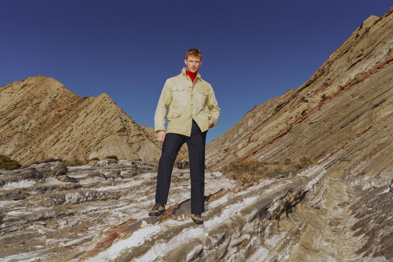 Man in a beige jacket and dark pants standing on rock formations in a desert landscape under a clear blue sky, full-length shot.