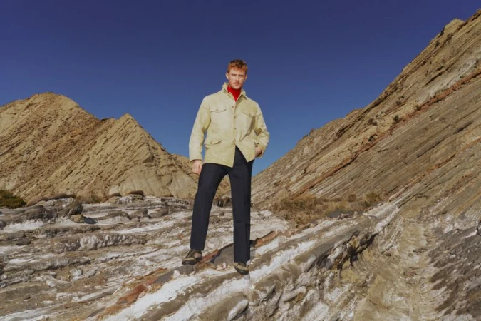 Man in a beige jacket and dark pants standing on rock formations in a desert landscape under a clear blue sky, full-length shot.
