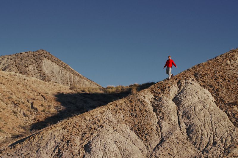 Adult walking on a ridge of a rugged, rocky, tan-colored mountain against a clear blue sky in a sunny outdoor environment.