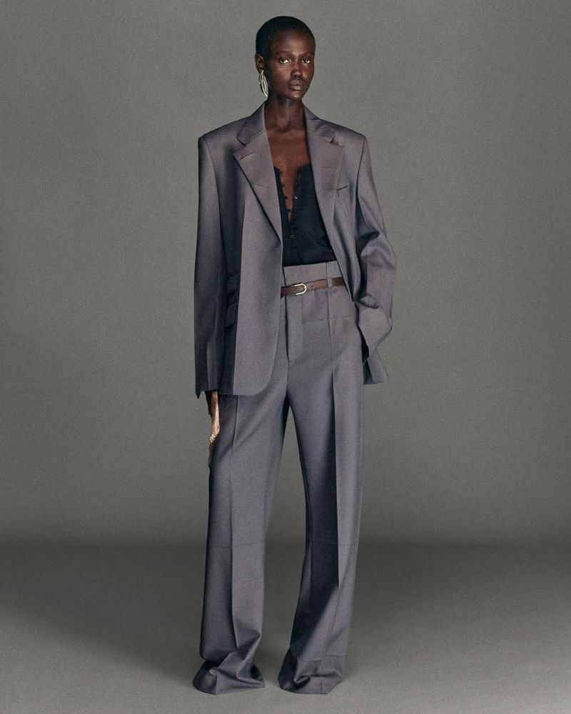 Fashion editorial shot of a Black woman posing in a gray oversized suit with wide leg pants on a neutral gray background. 