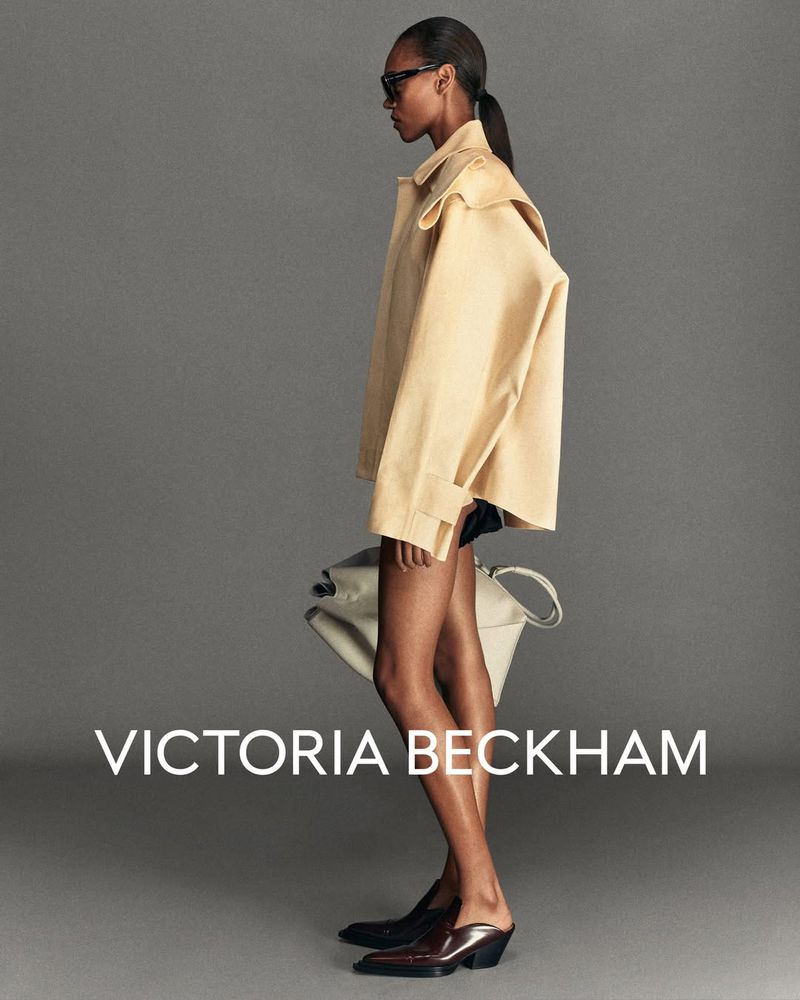 Fashion portrait: a slender Black woman models a beige jacket and slides, carrying a cream tote, against a gray studio backdrop.