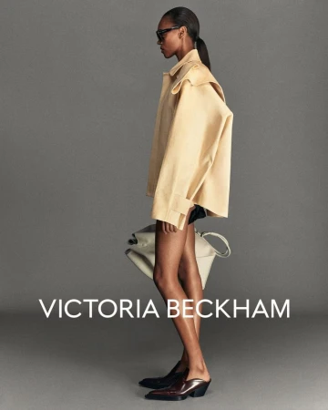 Fashion portrait: a slender Black woman models a beige jacket and slides, carrying a cream tote, against a gray studio backdrop.