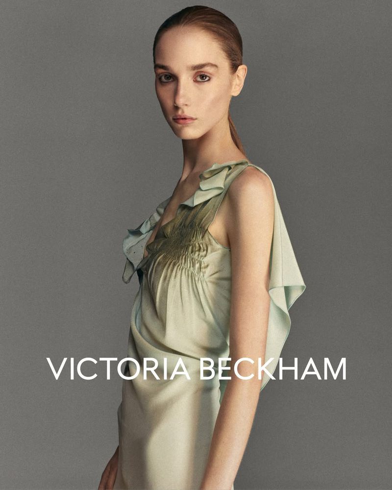 Fashion shot of a woman in a light green dress, posing in front of a grey backdrop with VICTORIA BECKHAM brand logo.