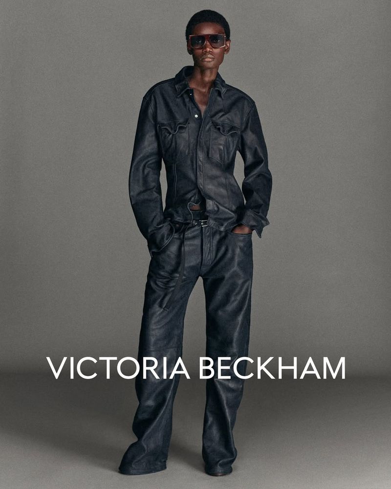 Fashion portrait of a black woman in a denim jumpsuit and sunglasses standing against a grey background in a studio.