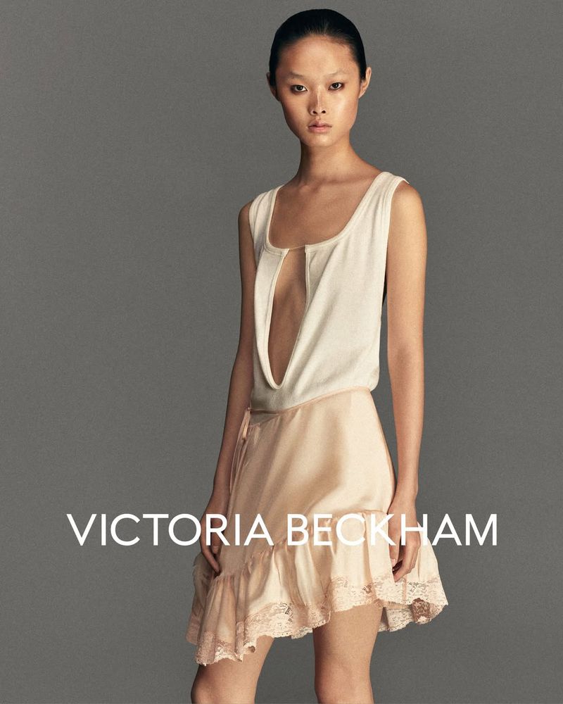 A woman with updo poses in front of grey background wearing a white sleeveless top with revealing front and pink layered skirt.