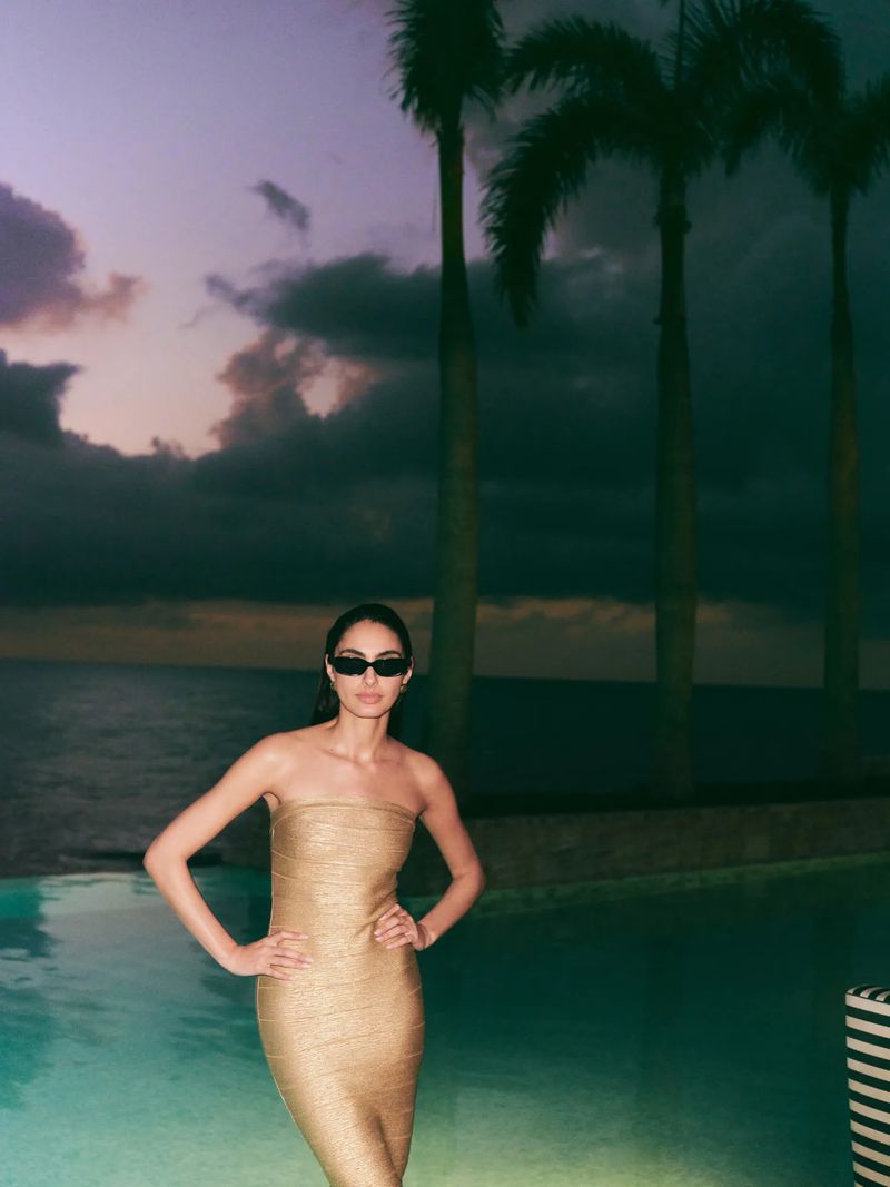 Woman in a gold dress and sunglasses poses near a pool with palm trees and a cloudy, colorful sky at dusk or dawn.