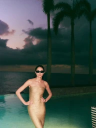 Model in strapless gold dress wearing black sunglasses standing poolside by ocean at sunset