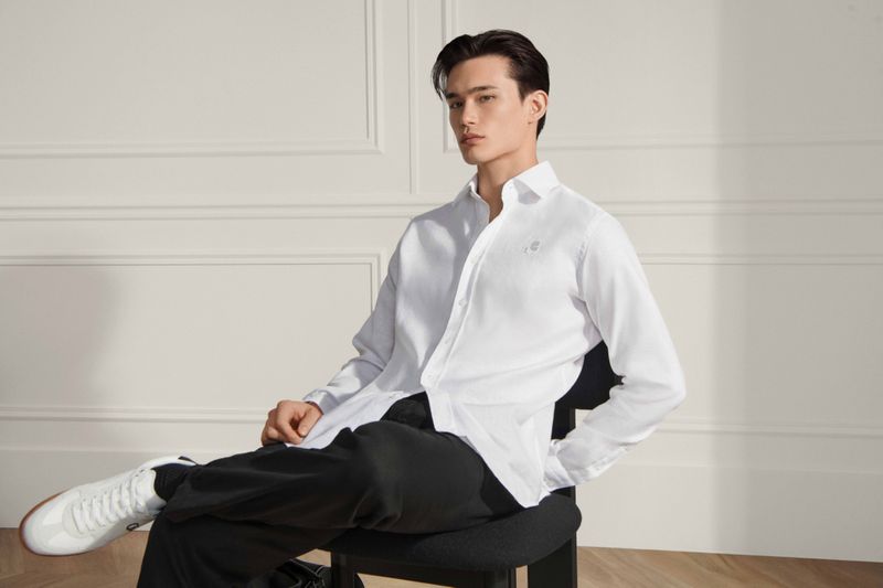 Indoor portrait of a young adult male in white shirt and black pants, seated on black chair in front of panelled wall.