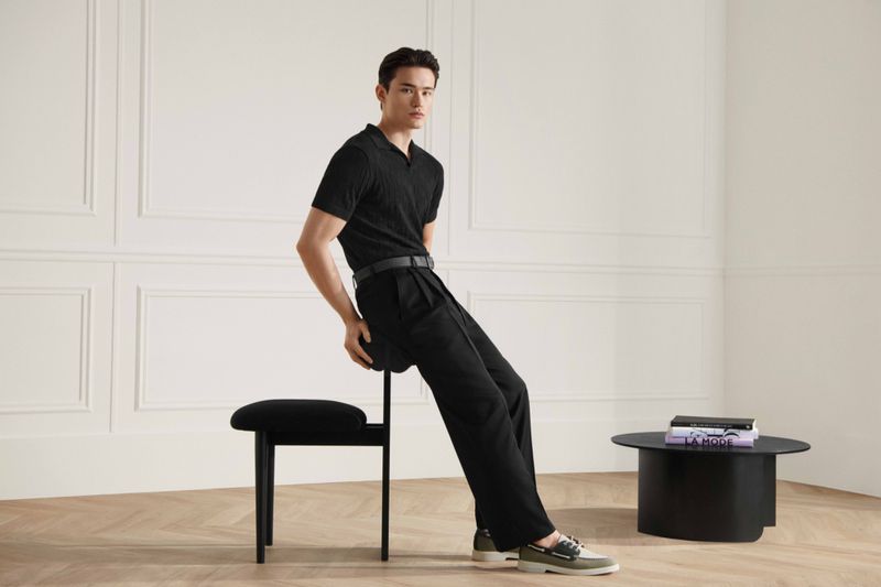 Fashion shot of a young adult male wearing a black polo shirt and black pants sitting on a black stool in a minimalist interior.