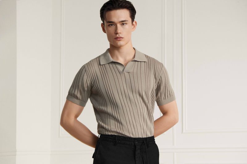 Adult male poses in a ribbed short-sleeved shirt and dark trousers against a neutral background. Fashion-forward with hands behind back.