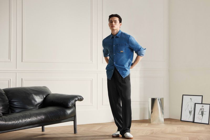 Man in blue denim shirt and black trousers stands in an indoor setting near black leather sofa and framed art prints.