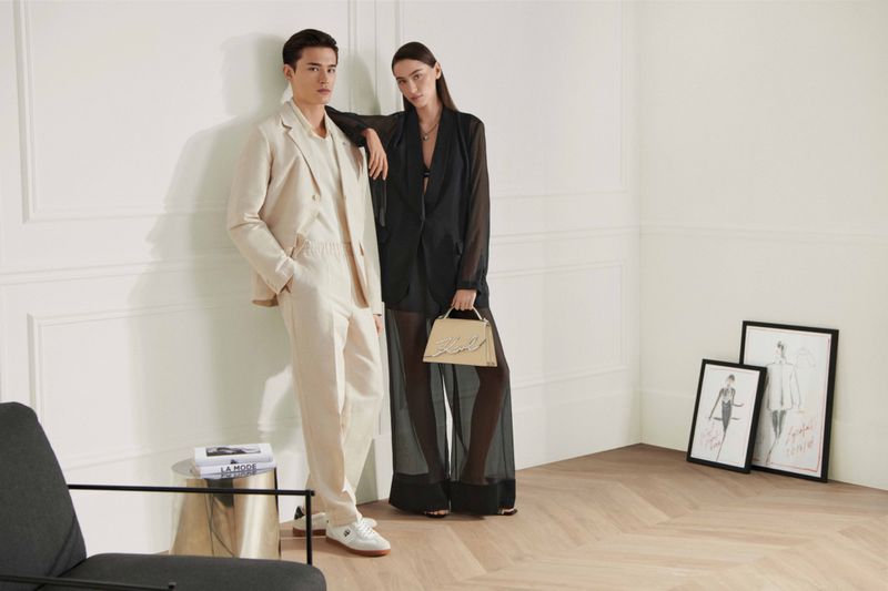 Fashionable couple strikes pose indoors. Man in cream suit, woman in sheer black top, bag in hand. Minimalist, clean backdrop with artwork.