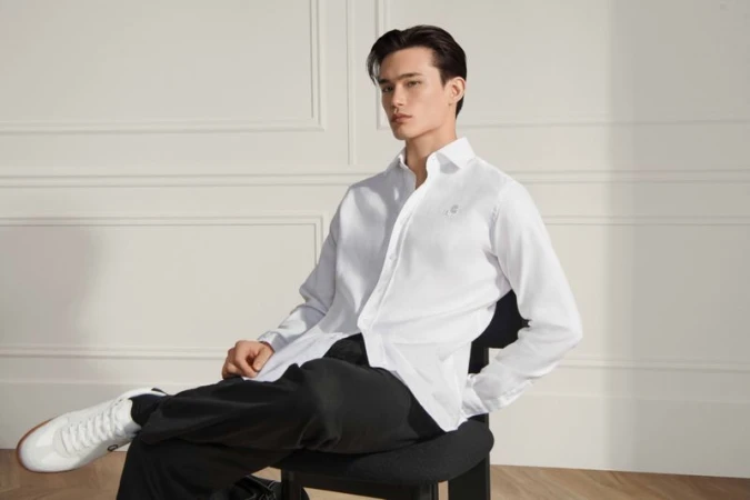 Model seated on a black chair wearing a white shirt and black trousers against a neutral wall with molding.