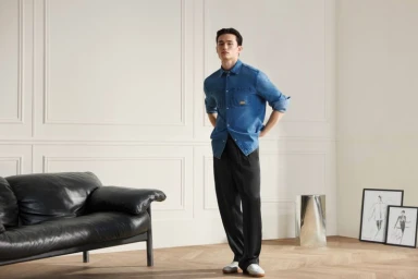 Model Alex Schlab in denim shirt and dark trousers standing in a minimalist room with a black leather sofa for Karl Lagerfeld SS26.