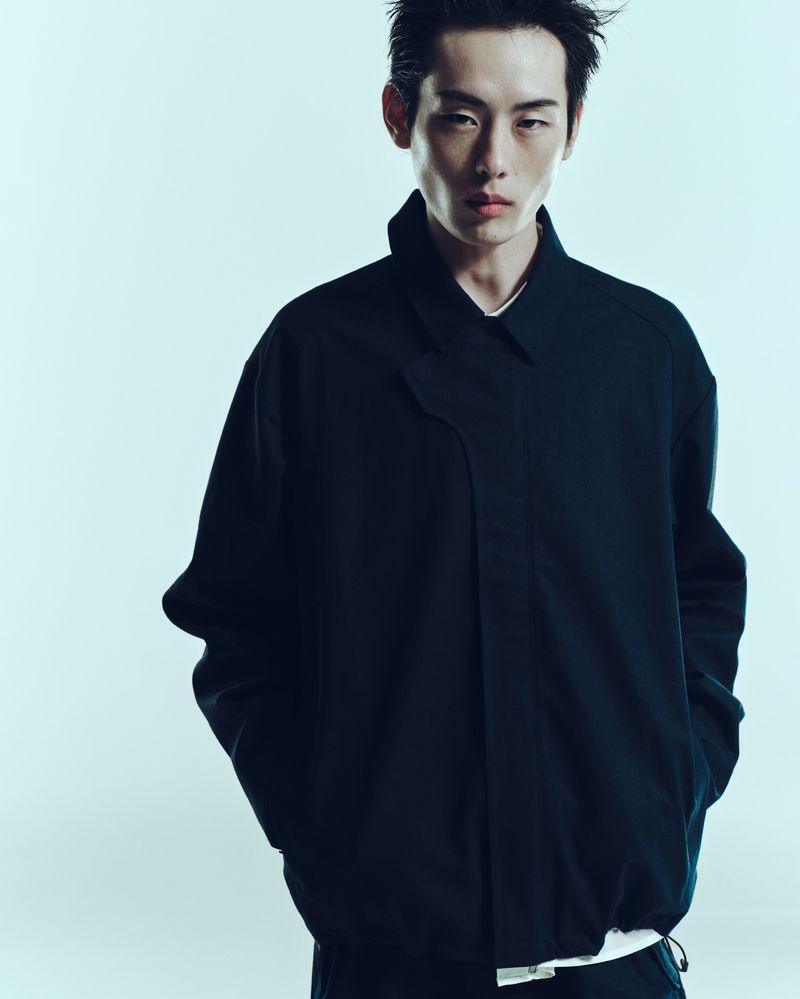Portrait of a slender Asian male model in a dark blue jacket against a graduated blue background in a clean studio fashion shot.