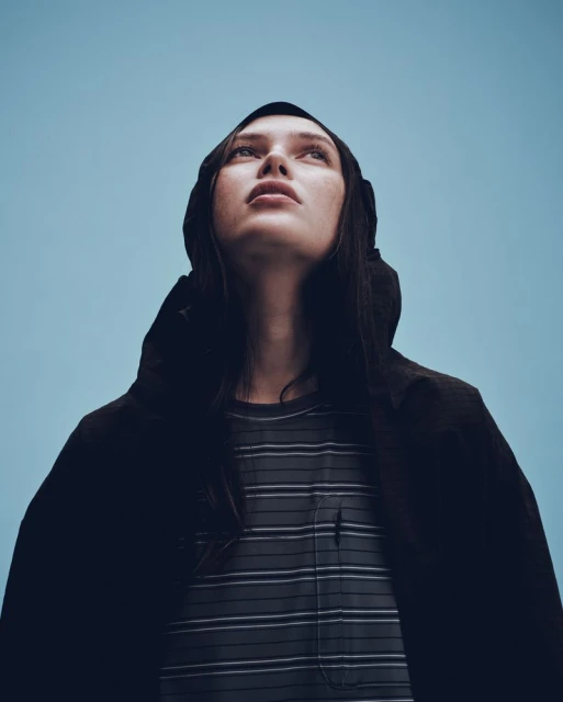 Low-angle portrait of a woman in a hooded coat looking up against a clear blue sky, highlighting fashion and atmospheric mood.