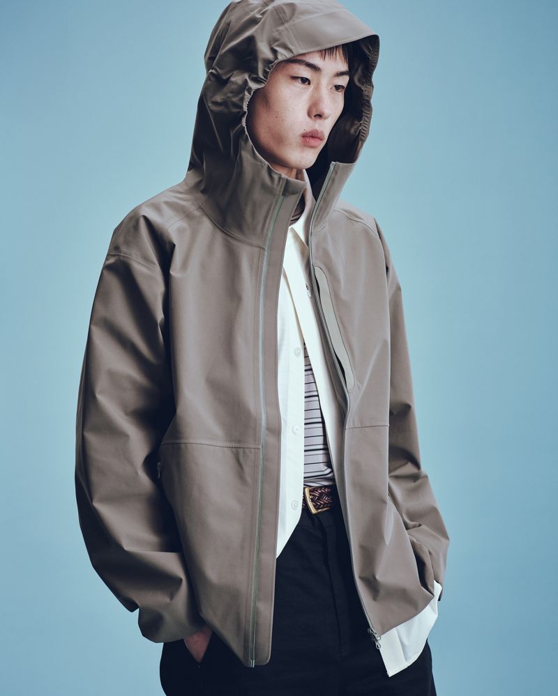 Adult male poses indoors wearing a taupe hooded jacket, a striped collared shirt, black pants, and a decorative belt against light blue.
