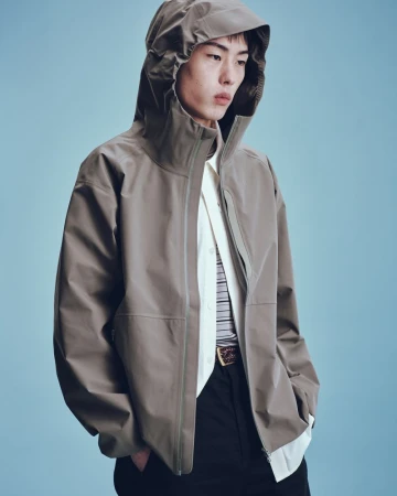 Adult male poses indoors wearing a taupe hooded jacket, a striped collared shirt, black pants, and a decorative belt against light blue.