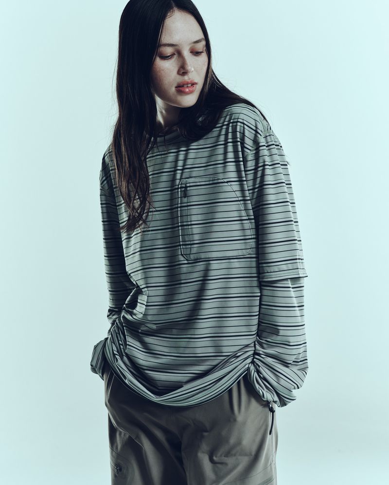 A young woman with long dark hair poses indoors wearing a striped long-sleeve shirt and brown pants in a fashion editorial shot.