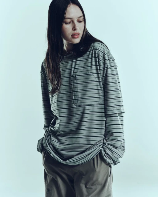 A young woman with long dark hair poses indoors wearing a striped long-sleeve shirt and brown pants in a fashion editorial shot.