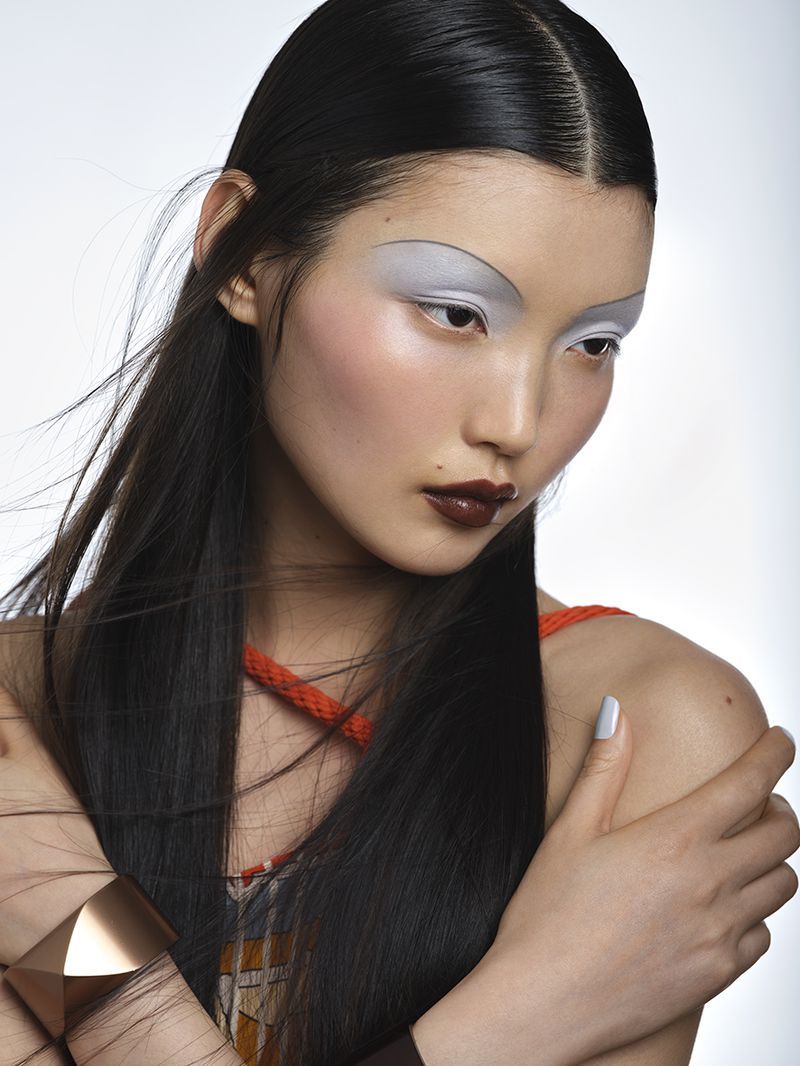 Portrait of an Asian woman with long dark hair, geometric gold bracelet, and unique grey and brown makeup against a white background.