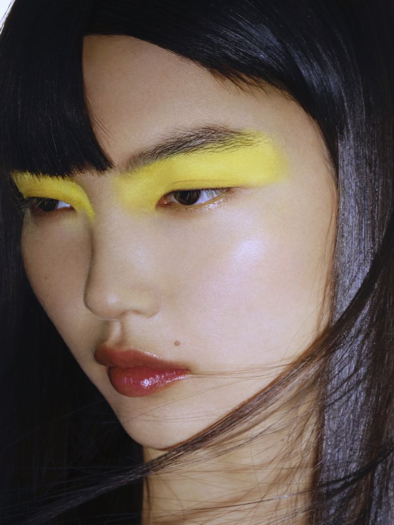 Close-up of Asian woman with yellow eyeshadow, dark hair, and glossy lips. Beauty portrait highlighting makeup and facial features.