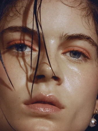 Close-up portrait of a young adult woman with wet hair, glowing skin, and bright eye makeup in a beauty fashion-style shot.