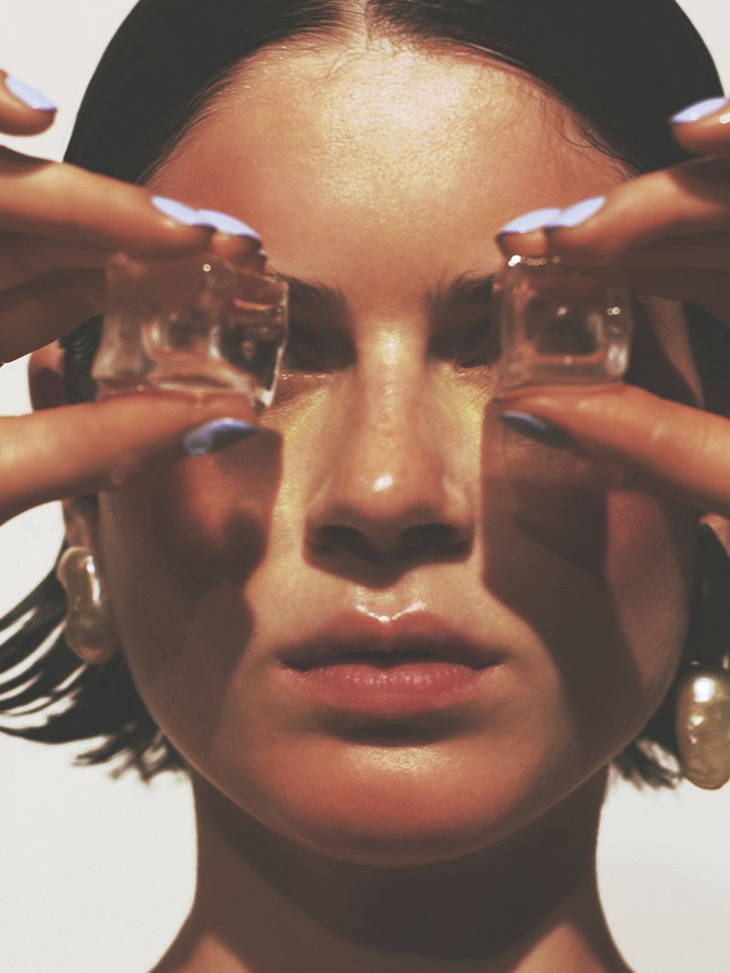 Close-up of a woman holding ice cubes to her eyes, showcasing pastel blue nails, light skin, and pearl earrings against a plain backdrop.