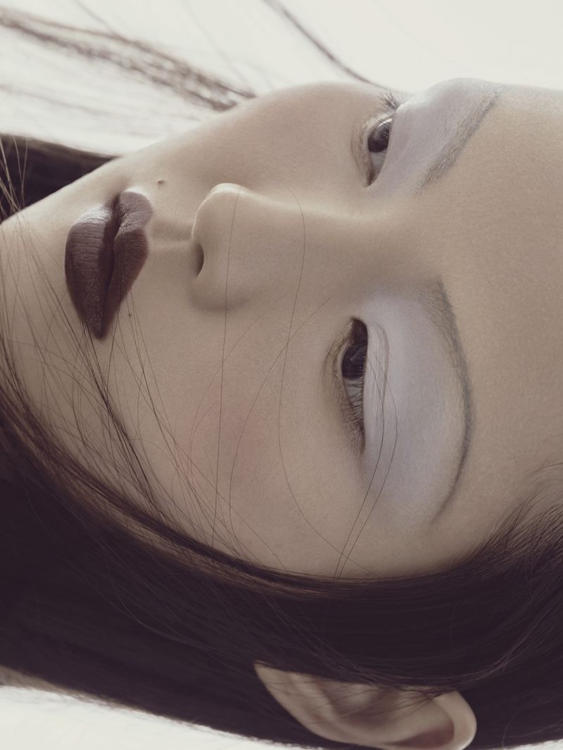 Close-up studio shot of a young Asian female model lying down, showcasing dark lipstick and light eyeshadow against a white backdrop.