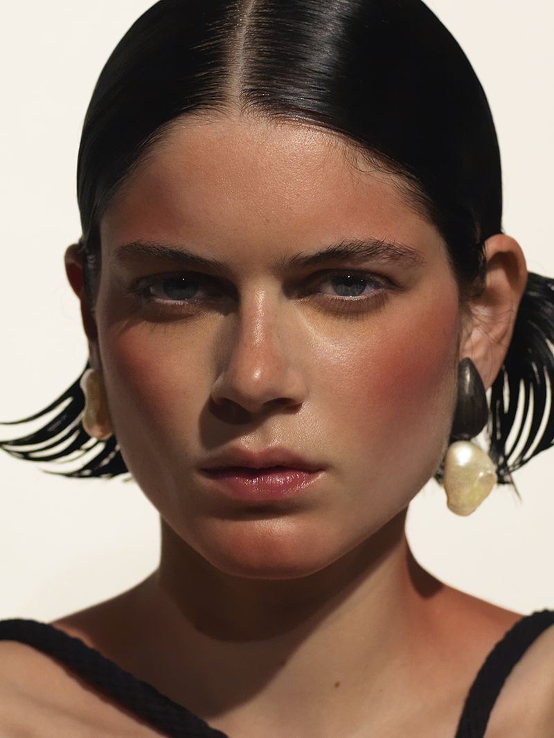 Close-up portrait of a young woman with glossy hair and dramatic rouge makeup, wearing statement earrings against a white backdrop.