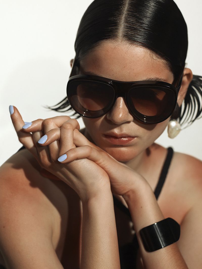 Close-up of a young adult woman with brown sunglasses, light-blue nail polish, and a black bracelet posing in front of a white background.