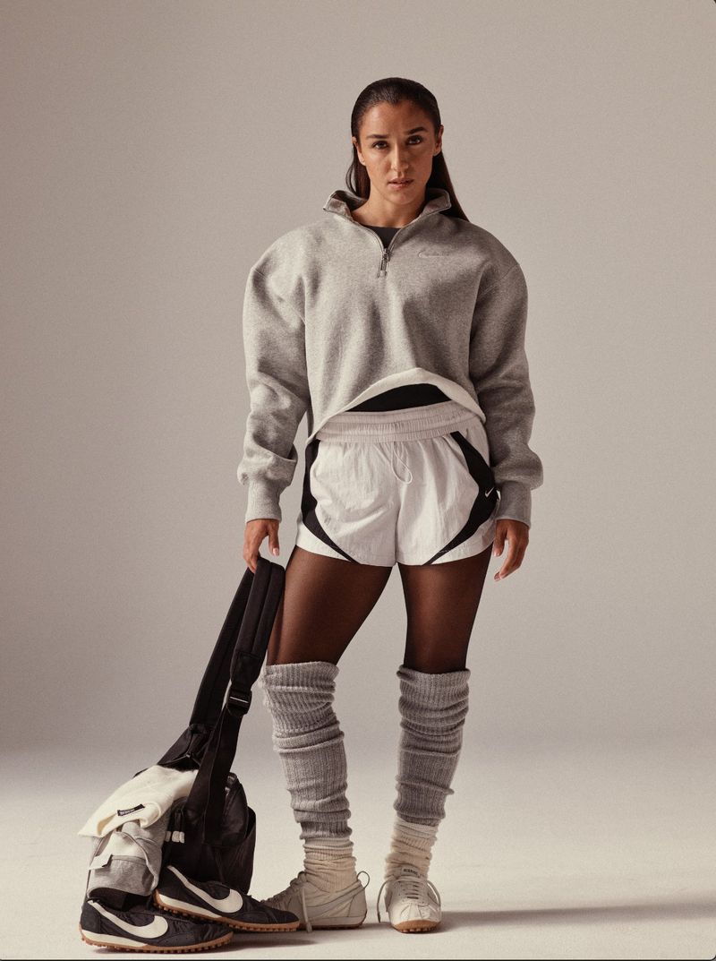 Athletic woman in grey sweatshirt and white shorts stands with gym bag and sneakers in a full studio shot, athletic wear fashion.