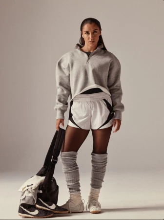 Athletic woman in grey sweatshirt and white shorts stands with gym bag and sneakers in a full studio shot, athletic wear fashion.