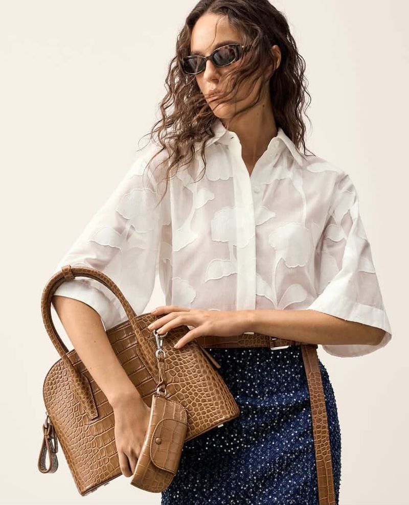 Adult female in white patterned top and blue skirt holding a brown textured handbag with an attached coin purse accessory.