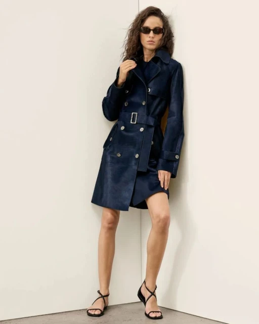 A slender woman poses indoors wearing a navy trench coat, a short skirt, black strappy sandals, and sunglasses against a neutral corner.