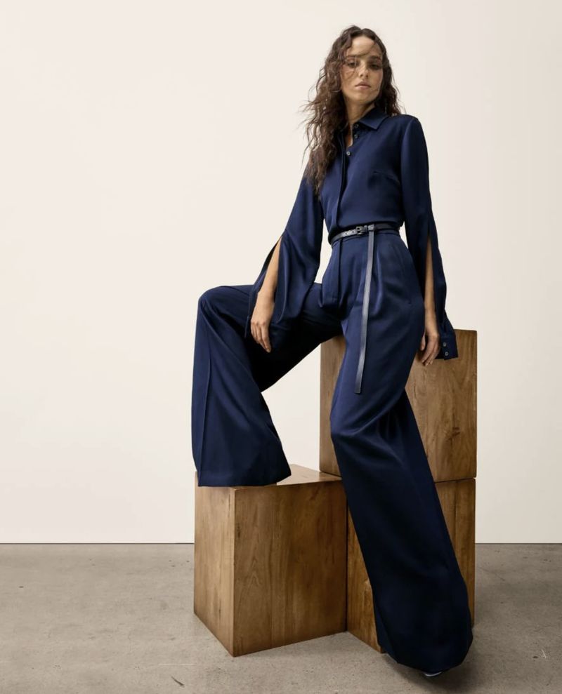 Brunette woman in fashionable navy-blue wide-leg pants and slit-sleeve top sitting on stacked wooden cubes in a minimalist studio setting.