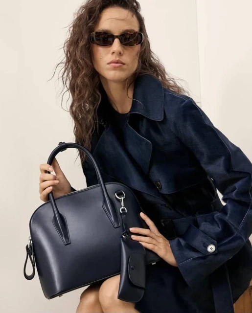 Elegant woman in a navy trench coat holding a sophisticated navy leather satchel bag, accessorized with sunglasses, in a studio setting.