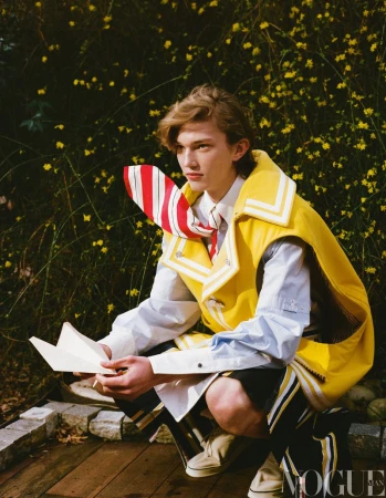Editorial fashion shot of a young male adult model in a yellow vest with a striped tie, holding a letter in an outdoor setting.