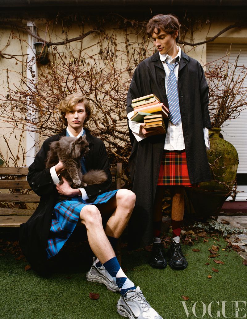 Two male fashion models outdoors wearing kilts and black coats, one holding books, the other holding a cat, with a brick wall backdrop.