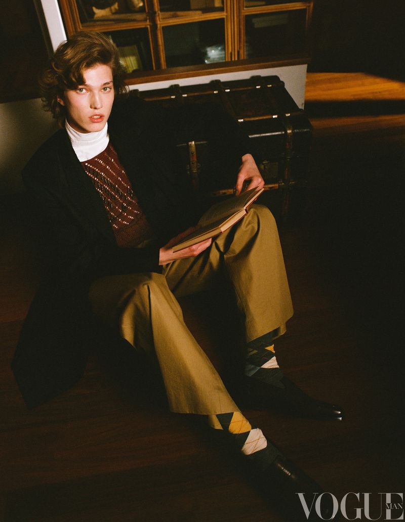 A young adult male is sitting and reading a book indoors near a trunk. He is wearing a dark blazer, brown pants, and patterned socks.